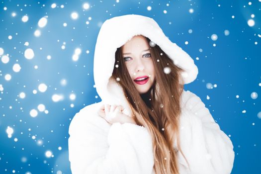 Happy Christmas and winter holiday portrait of young woman in white hooded fur coat, snow on blue background, fashion and lifestyle campaign