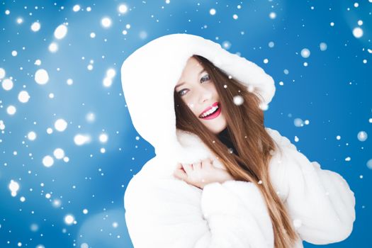 Happy Christmas and winter holiday portrait of young woman in white hooded fur coat, snow on blue background, fashion and lifestyle campaign