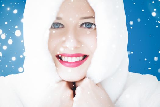 Happy Christmas and winter holiday portrait of young woman in white hooded fur coat, snow on blue background, fashion and lifestyle campaign