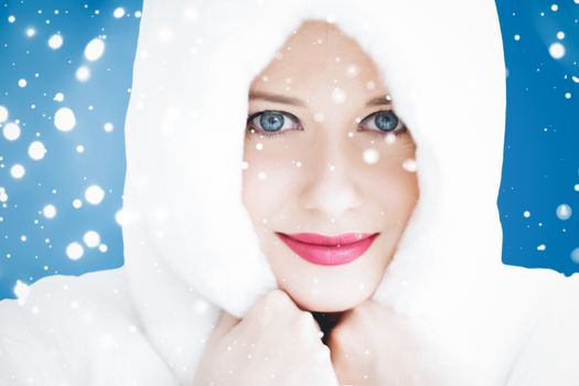 Happy Christmas and winter holiday portrait of young woman in white hooded fur coat, snow on blue background, fashion and lifestyle campaign