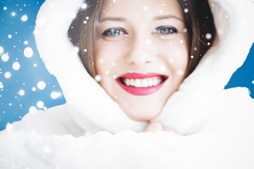 Happy Christmas and winter holiday portrait of young woman in white hooded fur coat, snow on blue background, fashion and lifestyle campaign