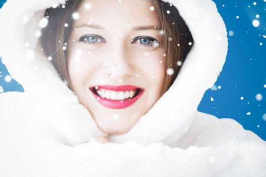 Happy Christmas and winter holiday portrait of young woman in white hooded fur coat, snow on blue background, fashion and lifestyle campaign