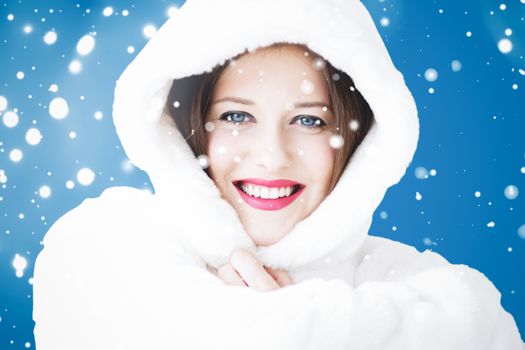 Happy Christmas and winter holiday portrait of young woman in white hooded fur coat, snow on blue background, fashion and lifestyle campaign