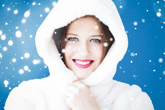 Happy Christmas and winter holiday portrait of young woman in white hooded fur coat, snow on blue background, fashion and lifestyle campaign