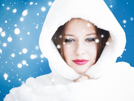 Happy Christmas and winter holiday portrait of young woman in white hooded fur coat, snow on blue background, fashion and lifestyle campaign