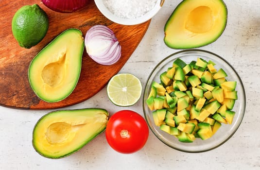 Avocado halves, some cut to small pieces, limes, tomato and onion - basic guacamole ingredients on white working board, view from above.