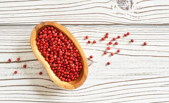 Red or Pink peppercorn in small wooden bowl, some scattered on white boards desk, closeup photo from above.