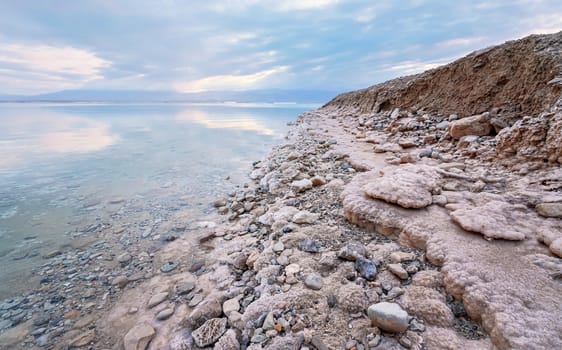 Sand and stones covered with crystalline salt on shore of Dead Sea, clear water near - typical scenery at Ein Bokek beach, Israel.