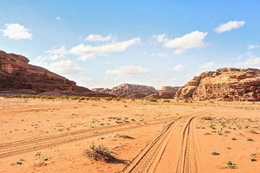 Rocky massifs on red sand desert, vehicle tracks ground, bright cloudy sky in background, typical scenery in Wadi Rum, Jordan.