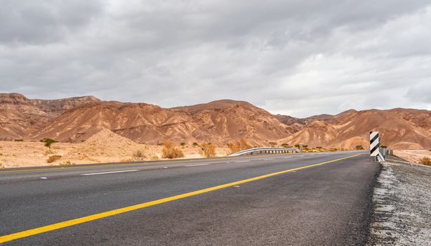 Modern asphalt highway leading through desert landscape in Southern region of Israel, cloudy sky above.