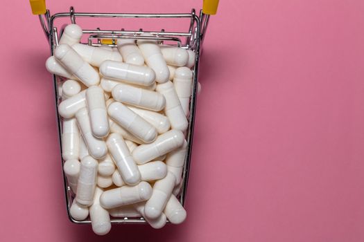 Top close up shot of a small shopping cart full of white pills. Pink background, copy space. Shopping online, buying medicine, pharmaceutical business concepts.