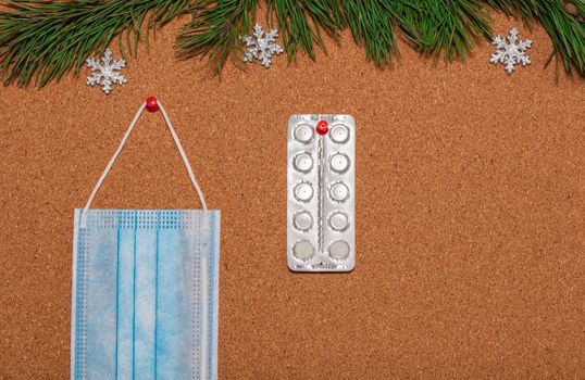 Medical mask and white pills pinned on a corkwood notice board which is decorated with pine twigs and silver snowflakes at the top. Healthcare, christmas, new years celebration, new normal concepts.