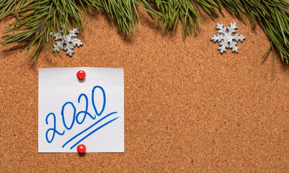 White sticky note with 2020 handwritten on it pinned on notice board which is decorated with pine twigs and snowflakes. 2020 Christmas, new years celebration season concept. Close-up shot. Copy space