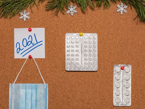 Medical mask, white pills, white sticky note with 2021 pinned on notice board which is decorated with pine twigs and snowflakes. Healthcare, christmas, new years celebration, new normal concepts.