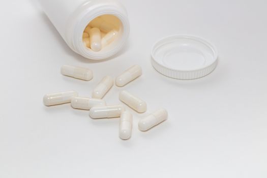 Bunch of white scattered pills on white background. White pills container and cap next to them slightly out of focus in the background. Pharmaceutical business and medicine sale concepts.