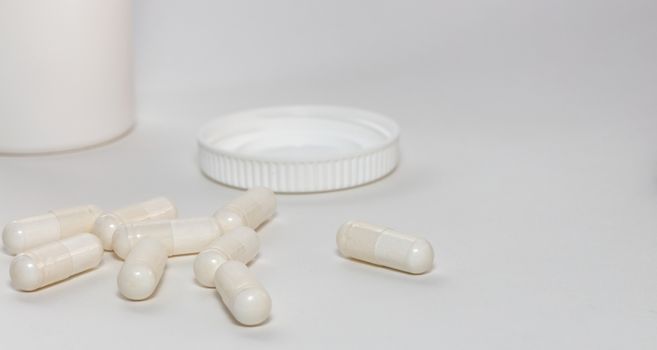 Bunch of white scattered pills on white background. White pills container and cap next to them slightly out of focus in the background. Close up. Pharmaceutical business and medicine sale concepts.