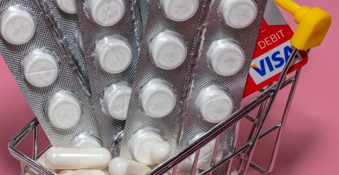 Barnaul, Russia - October 13, 2020: Close up shot of shopping cart with red visa debit card, white pills inside. Some out of focus. Pink background. Banner size. Pharmaceutical, shopping concepts