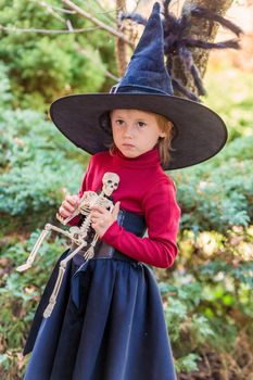 Little girl in a witch costume holding a skeleton on a halloween party in the garden