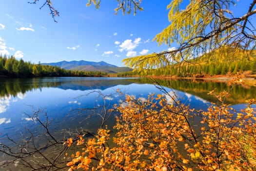 A beautiful flat surface of the lake against the background of colored hills and the blue sky. Fascinating view of the forest lake