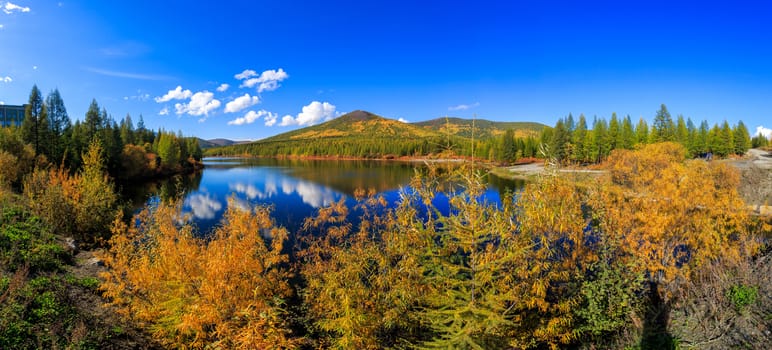 The nature of the Magadan region. Panoramic shot. A beautiful flat surface of the lake against the background of colored hills and the blue sky. Fascinating view of the forest lake