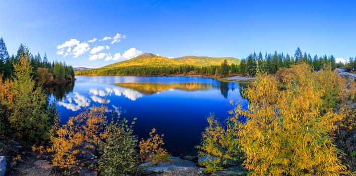 The nature of the Magadan region. Panoramic shot. A beautiful flat surface of the lake against the background of colored hills and the blue sky. Fascinating view of the forest lake