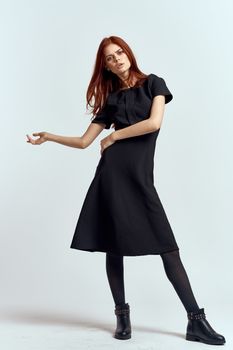 A woman in a black dress on a light background and pantyhose shoes red hair and pose in full growth. High quality photo