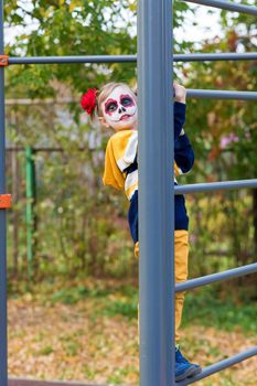 A little preschool girl with Painted Face, climbed the Swedish wall in the playground, celebrates Halloween or Mexican Day of the Dead.