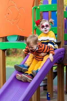 A little preschool girl with Painted Face, rides down the slide with her brother on the playground, celebrates Halloween or Mexican Day of the Dead.