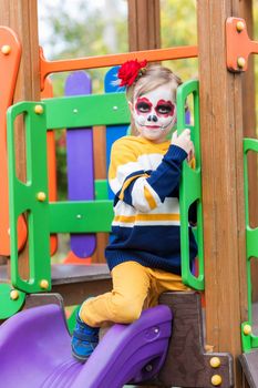 A little preschool girl with Painted Face, rides a slide on the playground, celebrates Halloween or Mexican Day of the Dead.