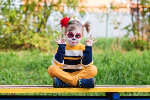 A little preschool girl with Painted Face, sitting on a bench in lotus position on the playground, celebrates Halloween or Mexican Day of the Dead.