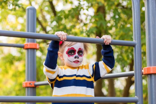 A little preschool girl with Painted Face, looking at the camera on the playground, celebrates Halloween or Mexican Day of the Dead..
