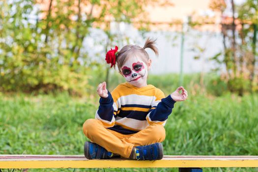 .A little preschool girl with Painted Face, smiling at the camera on the playground, celebrates Halloween or Mexican Day of the Dead.