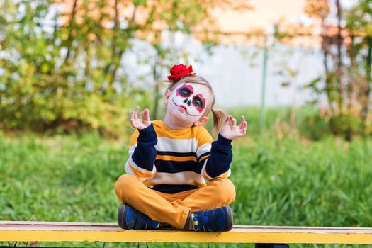 .A little preschool girl with Painted Face, smiling at the camera on the playground, celebrates Halloween or Mexican Day of the Dead.