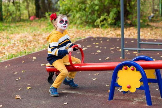 A little preschool girl with Painted Face, smiling at the camera on the playground, celebrates Halloween or Mexican Day of the Dead..