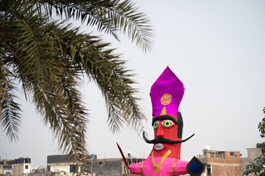 Huge colorful ravana effigy with a sword and a shield placed on the side of a busy street with a tree in the foreground and traffic passing by. Shows the preparations for the hindu festival of dussera prior to diwali celebrating the victory of good over evil as given in the hindu poem epic ramayana