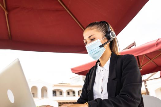Internet freelance job choice concept: a young cute woman with wireless headphones works on her laptop wearing a medical mask on a table of a bar - New normal jobs with remote connections and Covid 19