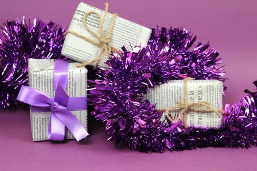 Gifts wrapped in old newspaper with purple bow, tinsel and rope on purple background