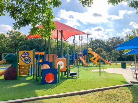 Modern neighborhood playground with sun shade sails, artificial grass in Flower Mound, Texas, America. Suburban recreational facility surrounded by wooden fence and tall trees