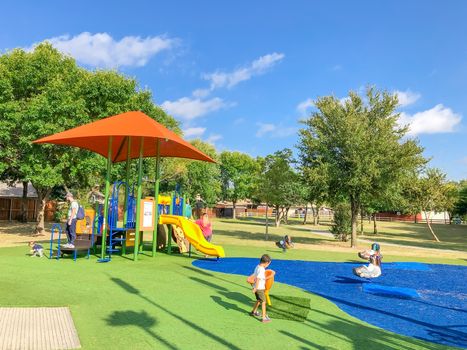 Large neighborhood playground with sun shade sails, artificial grass and kids playing in Flower Mound, Texas, America. Suburban recreational facility surrounded by wooden fence