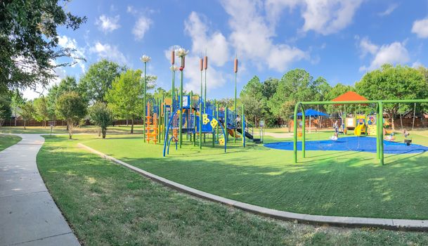 Panorama view neighborhood playground with sun shade sails, artificial grass in Flower Mound, Texas, America. Suburban recreational facility surrounded by wooden fence and large mature trees