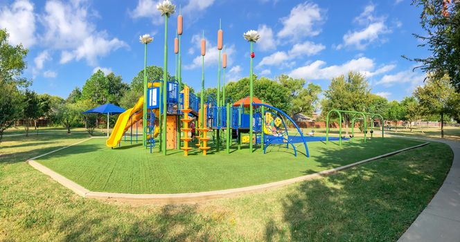Panorama view neighborhood playground with sun shade sails, artificial grass in Flower Mound, Texas, America. Suburban recreational facility surrounded by wooden fence and large mature trees