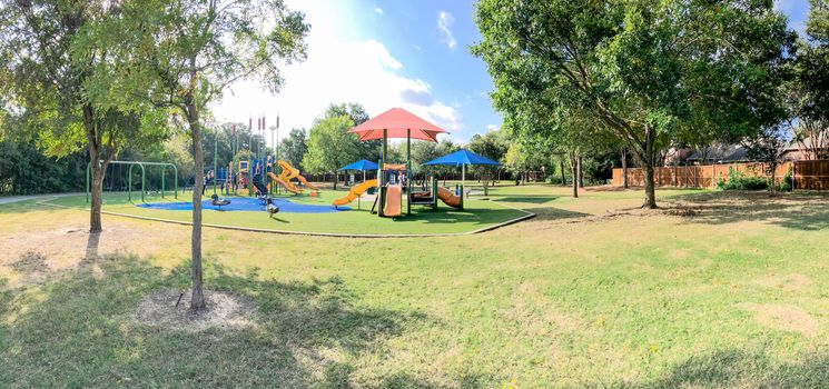 Panorama view neighborhood playground with sun shade sails, artificial grass in Flower Mound, Texas, America. Suburban recreational facility surrounded by wooden fence and large mature trees