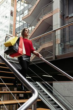 Adult woman wearing a red sweater walking down the stairs of a shopping mall carrying colorful shopping bags.