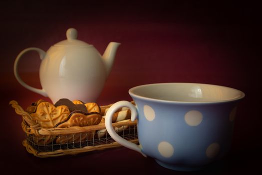 White ceramic kettle, cookies and a blue cup on a maroon background