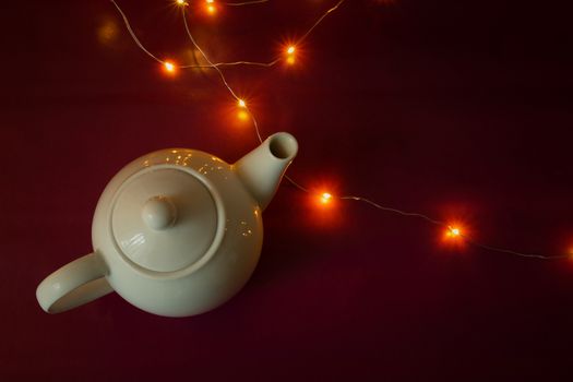 White ceramic kettle and Christmas lights on burgundy background, top view