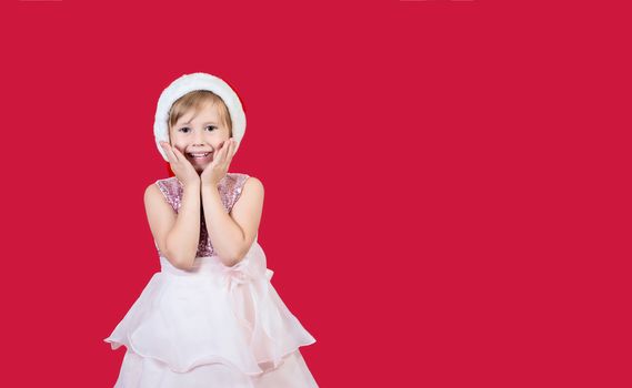 Surprised funny cacusian cute child girl in santa hat looking at the camera waiting for presents celebrating happy 2021 New Year isolated on red background. Merry Christmas presents shopping sale.
