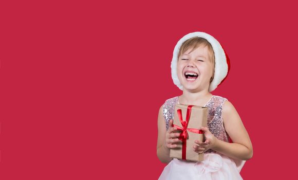 Surprised funny cacusian cute child girl in santa hat looking at the camera, smiling widly happily and hugs a gift isolated on red background. Merry Christmas presents shopping sale.