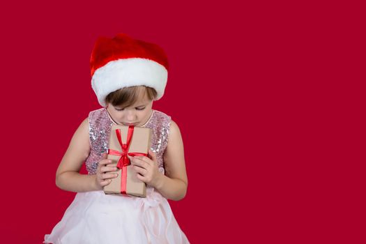 Beautiful cute cacusian little girl in santa hat holding a present to her chest sad maybe her christmas is ruined isolated on red background.