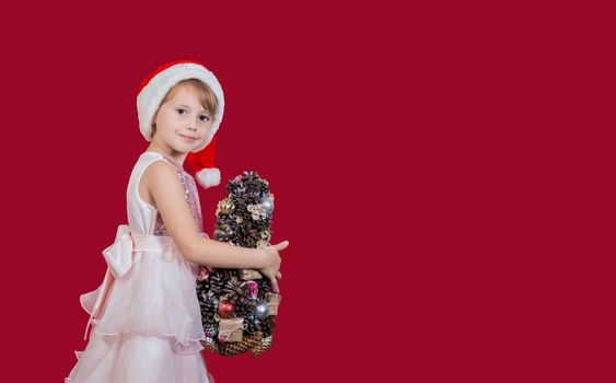Beautiful cute cacusian little girl in santa hat looking at the camera with homemade christmas tree celebrating happy 2021 New Year isolated on red background. Merry Christmas presents shopping sale.