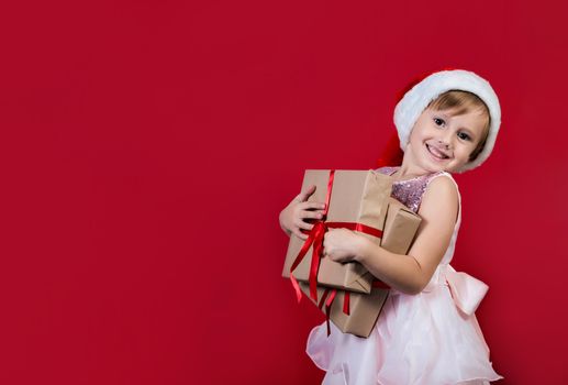 Excited funny cacusian cute child girl wearing santa hat looking at camera holding many gifts boxes celebrating happy 2021 New Year isolated on red background. Merry Christmas presents shopping sale.